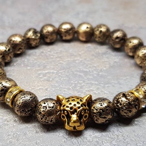 Könnte beinhalten: Ein goldfarbenes Armband mit einem strukturierten Lava-Stein-Perlenmuster und einem goldfarbenen Jaguar-Kopf-Anhänger.