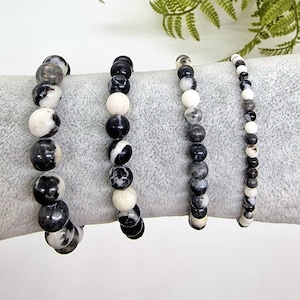 Puede incluir: Cuatro pulseras hechas de cuentas marmoleadas en blanco y negro. Las pulseras están apiladas sobre una superficie gris.