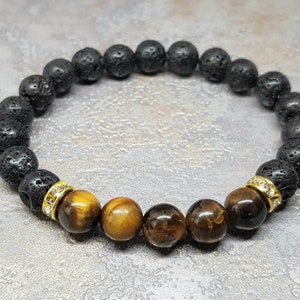 Puede incluir: Un brazalete de cuentas de lava negra y ojo de tigre con separadores dorados y detalles de cristal transparente.
