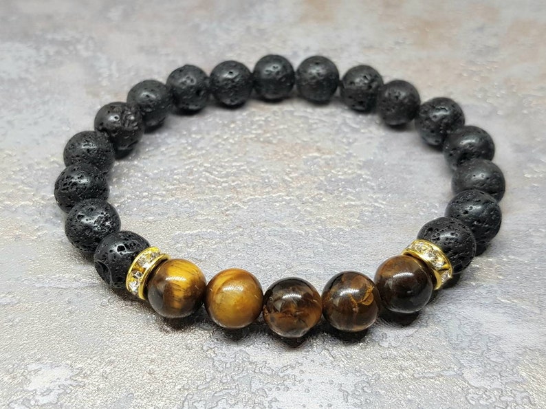 Puede incluir: Un brazalete de cuentas de lava negra y ojo de tigre con separadores dorados y detalles de cristal transparente.