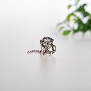 Peut inclure: Un charme en argent en forme de singe avec des yeux bleus et une chaîne attachée à sa queue. Le singe est recouvert de petits cristaux transparents.