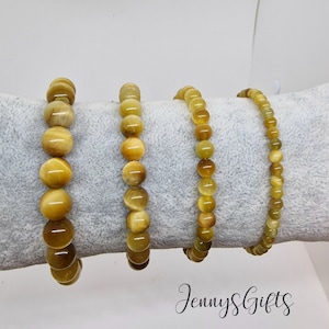 May include: Four bracelets made of yellow tiger's eye beads. The bracelets are arranged in a row on a gray fabric surface. The bracelets are all the same size and have a similar design. The text "Jenny's Gifts" is visible in the lower right corner of the image.
