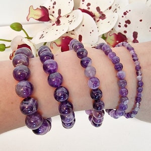 Peut inclure: Quatre bracelets en améthyste empilés sur un poignet. Les bracelets sont faits de perles rondes de différentes tailles et nuances de violet.