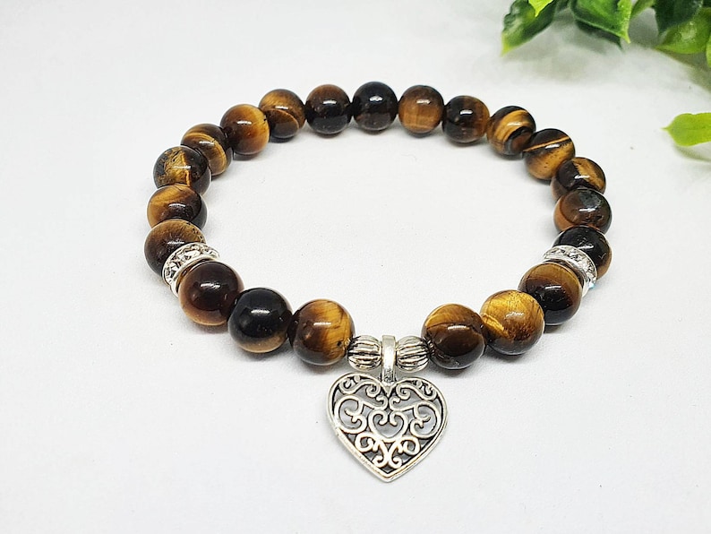 May include: A brown and gold tiger's eye beaded bracelet with a silver heart charm. The bracelet is made with round beads and has a silver clasp.