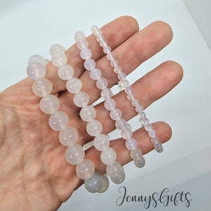 Może przedstawiać: Cztery białe bransoletki z kamieni szlachetnych w różnych rozmiarach, przy czym największa bransoletka ma największe koraliki. Bransoletki wykonane są z okrągłych, gładkich koralików.