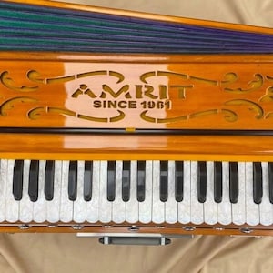 May include: A wooden harmonium with a polished finish, featuring a keyboard with white patterned keys and black keys. The top panel displays the text AMRIT SINCE 1961 with decorative cutouts. A purple and green bellows system is attached at the top.