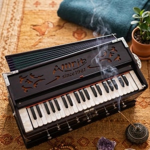 May include: A dark brown wooden harmonium featuring white and black keys, with the text "Amrit since 1961" engraved on the front panel. The instrument includes a bellows system, metal corner accents, and decorative cutouts, with smoke rising.