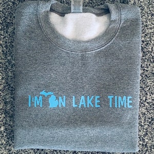 Puede incluir: Sudadera gris con un contorno azul del estado de Michigan y el texto "I'm on Lake Time".