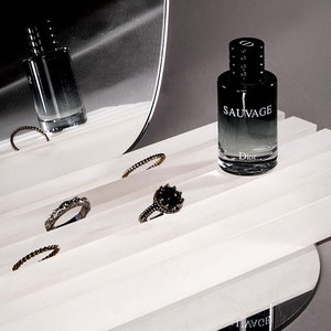 May include: A black glass bottle of Dior Sauvage cologne on a white platform with silver rings. The bottle has a black cap and a silver label with the words "Sauvage" and "Dior" printed on it.
