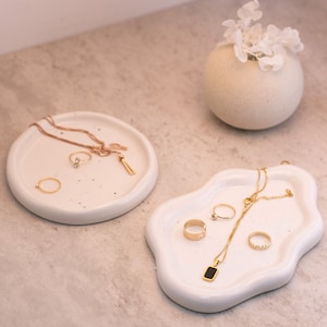 Op de afbeelding: Twee witte keramische schalen met gouden sieraden, waaronder kettingen, ringen en een hanger. De schalen staan op een lichtgrijze ondergrond.