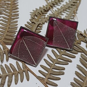 May include: A pair of square-shaped earrings with a dark purple resin background and a delicate white leaf pressed inside.
