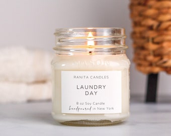 May include: A clear glass jar candle with a white label that reads "Ranita Candles Laundry Day 8 oz Soy Candle handpoured in New York". The candle is lit and the flame is visible.