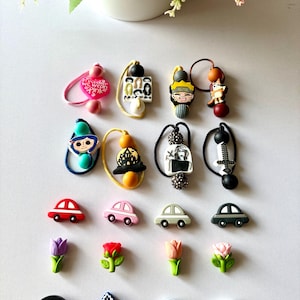 May include: A selection of decorative charms and accessories, including car-shaped charms in red, pink, grey, and black, and flower-shaped charms. Other charms feature cartoon characters and team logos. The charms are arranged on a white surface.