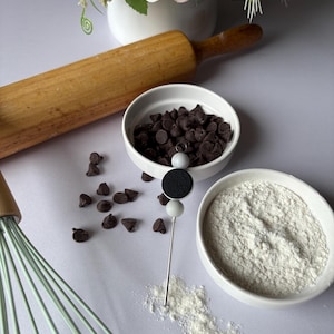 Peut inclure: Image sur le thème de la pâtisserie avec un rouleau à pâtisserie, un fouet et des bols de pépites de chocolat et de farine. Une épingle décorative avec un motif de biscuit est également présente, suggérant une mise au point sur la pâtisserie et les desserts.