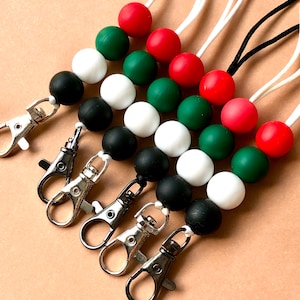 May include: A selection of lanyards featuring beads in red, green, white, and black. Each lanyard has a silver-coloured metal clasp. The beads are strung on white or black cords.