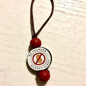 May include: A brown cord with two red beads attached to a white round tag with the text "NO PEANUTS ALLERGY ALERT" and a red circle with a diagonal line through a peanut image.