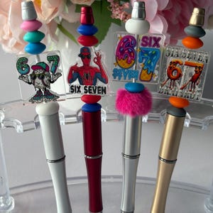 May include: A selection of pens with decorative toppers and charms. The pens have silver, gold, and red barrels. The toppers include stacked beads in various colours, a pink pom-pom, and charms with the numbers six and seven, and a Spider-Man graphic.
