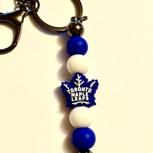 May include: A blue and white Toronto Maple Leafs keychain with a silicone bead charm. The keychain has a silver metal ring and a silver metal clasp.