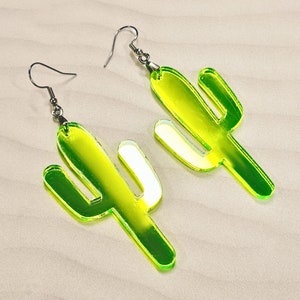May include: A pair of green cactus-shaped earrings with silver hooks. The earrings are made of clear acrylic and have a reflective finish.