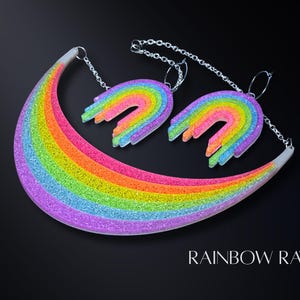 May include: A colorful necklace and matching earrings set with a rainbow design. The jewelry features a curved rainbow pendant and two rainbow-shaped earrings, all with glitter accents. The text "RAINBOW RAVE" is displayed at the bottom.