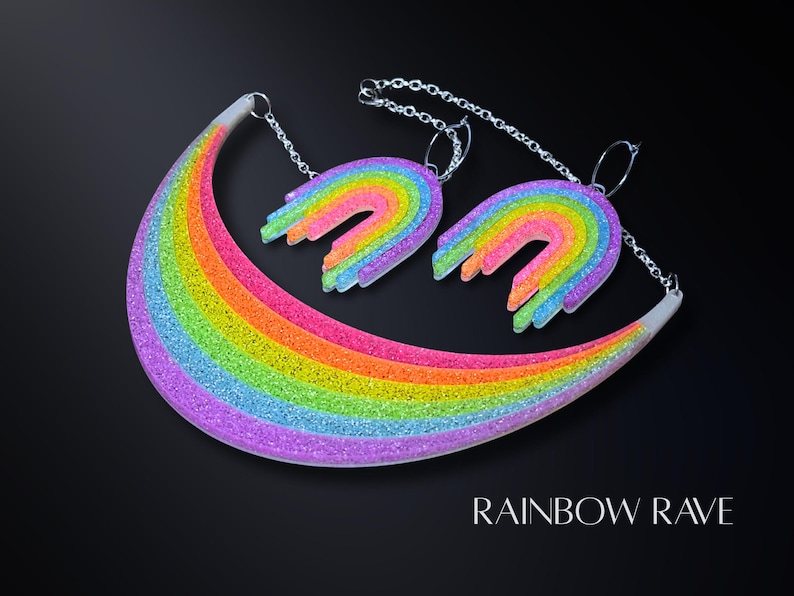 May include: A colorful necklace and matching earrings set with a rainbow design. The jewelry features a curved rainbow pendant and two rainbow-shaped earrings, all with glitter accents. The text "RAINBOW RAVE" is displayed at the bottom.