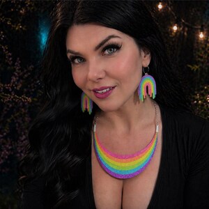 May include: A woman wearing a black top, rainbow-colored necklace, and matching rainbow earrings. The necklace and earrings are in the shape of a rainbow with a dripping effect. The background is dark with soft lighting.