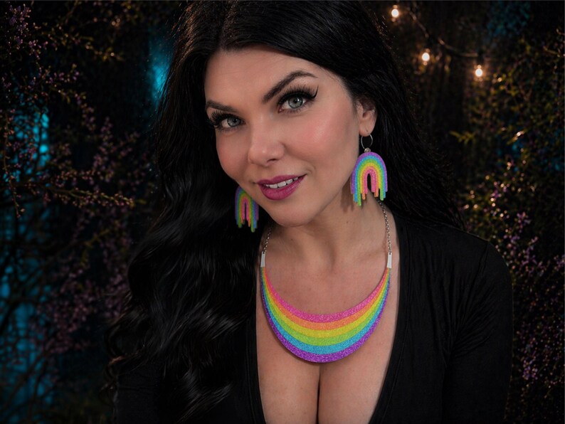 May include: A woman wearing a black top, rainbow-colored necklace, and matching rainbow earrings. The necklace and earrings are in the shape of a rainbow with a dripping effect. The background is dark with soft lighting.