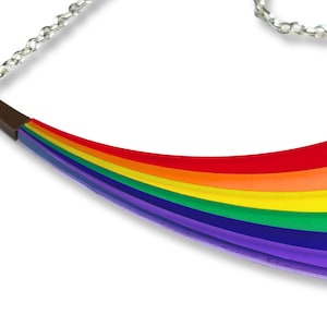 May include: A rainbow-colored necklace with a silver chain. The pendant is curved, with a brown metal accent at the top. The rainbow design features bands of red, orange, yellow, green, blue, and purple.