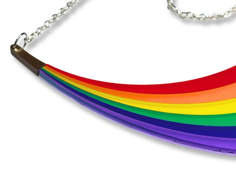 May include: A rainbow-colored necklace with a silver chain. The pendant is curved, with a brown metal accent at the top. The rainbow design features bands of red, orange, yellow, green, blue, and purple.