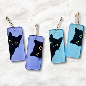 May include: Four rectangular earrings with a black cat design. The cats peek from the side of the earrings, with gold hoops. Two earrings are light blue, and two are purple. The cats have yellow or green eyes.