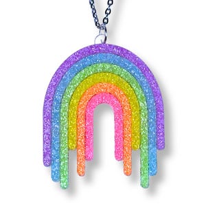 Op de afbeelding: Een regenboogvormige hanger aan een ketting met een veelkleurige glitterafwerking. Het ontwerp bestaat uit concentrische bogen in paars, blauw, groen, geel, oranje en roze, waarbij elke kleurband in een druipeffect naar beneden loopt aan een zilveren ketting.