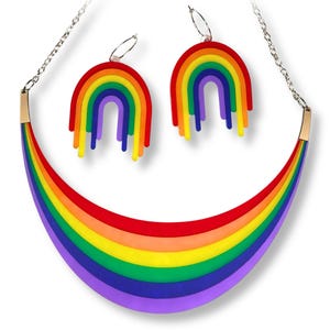 May include: A rainbow-themed jewelry set featuring a necklace and matching earrings. The necklace has a curved, layered design in red, orange, yellow, green, blue, and purple. The earrings are rainbow-shaped with dangling stripes. The set is on a white background.