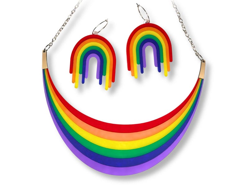 May include: A rainbow-themed jewelry set featuring a necklace and matching earrings. The necklace has a curved, layered design in red, orange, yellow, green, blue, and purple. The earrings are rainbow-shaped with dangling stripes. The set is on a white background.