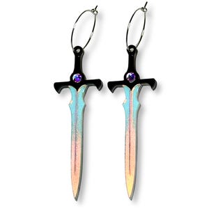 May include: A pair of hoop earrings featuring sword-shaped pendants. The sword hilts are black with a small, round, iridescent purple gem at the center. The blades transition from a light blue at the top to a warm peach and gold gradient at the tip.