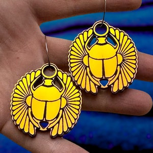 Op de afbeelding: Een paar gouden oorbellen met een scarabäus-ontwerp. De oorbellen zijn iriserend en hebben een gedetailleerd ontwerp.