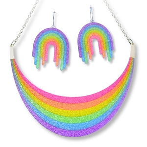 May include: A rainbow-themed jewelry set featuring a necklace and matching earrings. The necklace is a curved rainbow design with a silver chain. The earrings are rainbow-colored hoops. The jewelry has a glittery finish and is set against a white background.