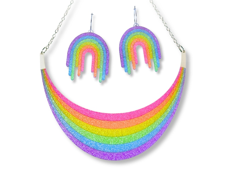 May include: A rainbow-themed jewelry set featuring a necklace and matching earrings. The necklace is a curved rainbow design with a silver chain. The earrings are rainbow-colored hoops. The jewelry has a glittery finish and is set against a white background.