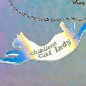 May include: A silver chain necklace with a clear iridescent pendant shaped like a cat. The pendant has the text "childless cat lady" printed on it.