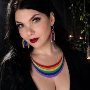 May include: A rainbow-themed jewelry set featuring a necklace and matching earrings. The necklace is a curved design with layers of red, orange, yellow, green, blue, and purple. The earrings are small rainbow arches. The jewelry is worn.
