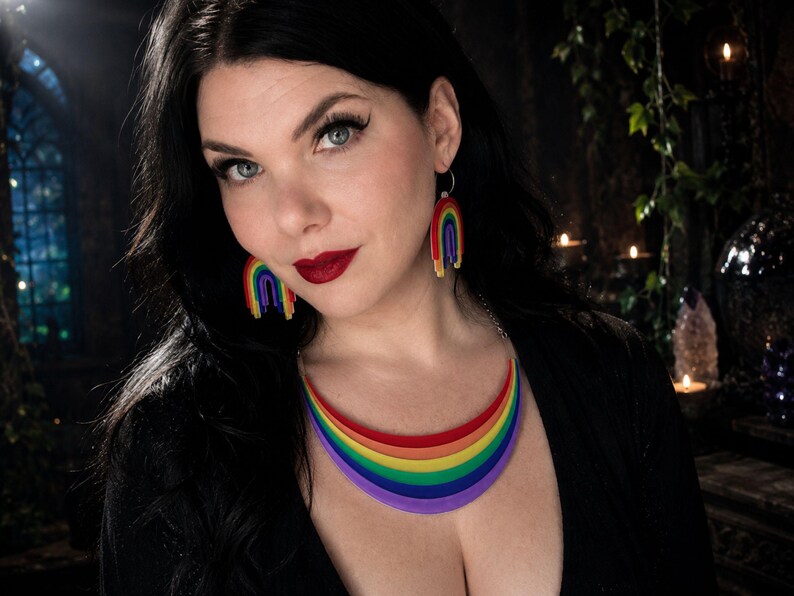 May include: A rainbow-themed jewelry set featuring a necklace and matching earrings. The necklace is a curved design with layers of red, orange, yellow, green, blue, and purple. The earrings are small rainbow arches. The jewelry is worn.