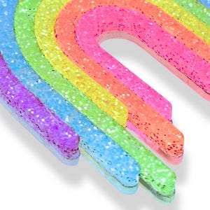 May include: A colorful rainbow-shaped decoration with glitter. The rainbow features layers of purple, blue, green, yellow, orange, and pink. The glitter adds a sparkling effect to the vibrant colors. The decoration is likely for crafts or decor.