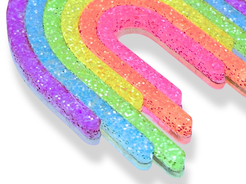 May include: A colorful rainbow-shaped decoration with glitter. The rainbow features layers of purple, blue, green, yellow, orange, and pink. The glitter adds a sparkling effect to the vibrant colors. The decoration is likely for crafts or decor.