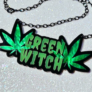 Puede incluir: Un collar de cadena negra con un colgante verde y negro. El colgante presenta un diseño de hoja de marihuana y las palabras "GREEN WITCH".