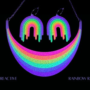May include: A UV reactive rainbow necklace and matching earrings set. The jewelry features a vibrant rainbow design with dripping accents. The necklace and earrings glow brightly in neon colors against a black background. The text "UV REACTIVE" and "RAINBOW RAVE" are visible.