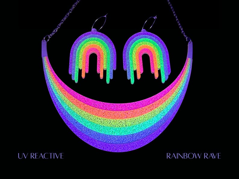 May include: A UV reactive rainbow necklace and matching earrings set. The jewelry features a vibrant rainbow design with dripping accents. The necklace and earrings glow brightly in neon colors against a black background. The text "UV REACTIVE" and "RAINBOW RAVE" are visible.