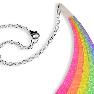 May include: A colorful rainbow-shaped necklace with a silver chain and clasp. The rainbow features layers of pink, orange, yellow, green, blue, and purple glitter. The necklace is designed to be worn around the neck.