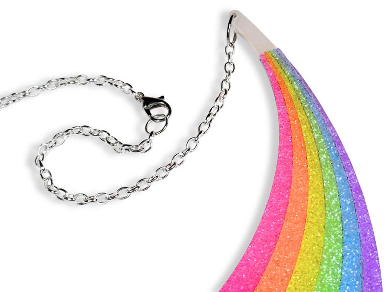 May include: A colorful rainbow-shaped necklace with a silver chain and clasp. The rainbow features layers of pink, orange, yellow, green, blue, and purple glitter. The necklace is designed to be worn around the neck.