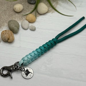 May include: A teal and light blue ombre paracord keychain with a silver metal charm featuring a manta ray silhouette. The keychain is on a white surface with small rocks.