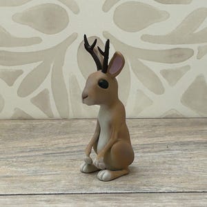 May include: A quirky jackalope figurine, a mythical animal combining a rabbit's body with antlers. The jackalope is mainly tan with white details on its paws and pink inner ears. The antlers are dark brown.