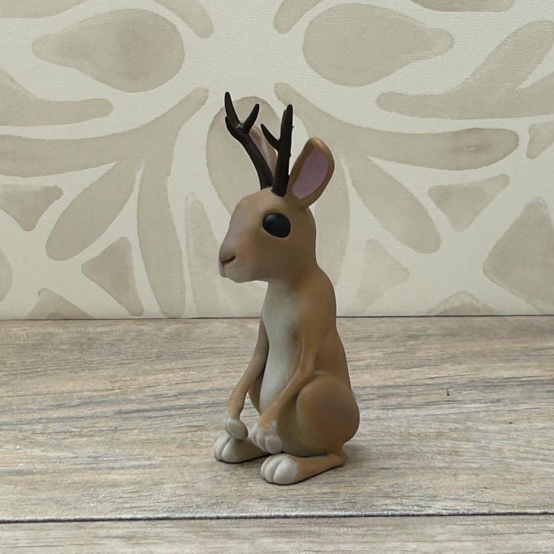 May include: A quirky jackalope figurine, a mythical animal combining a rabbit's body with antlers. The jackalope is mainly tan with white details on its paws and pink inner ears. The antlers are dark brown.